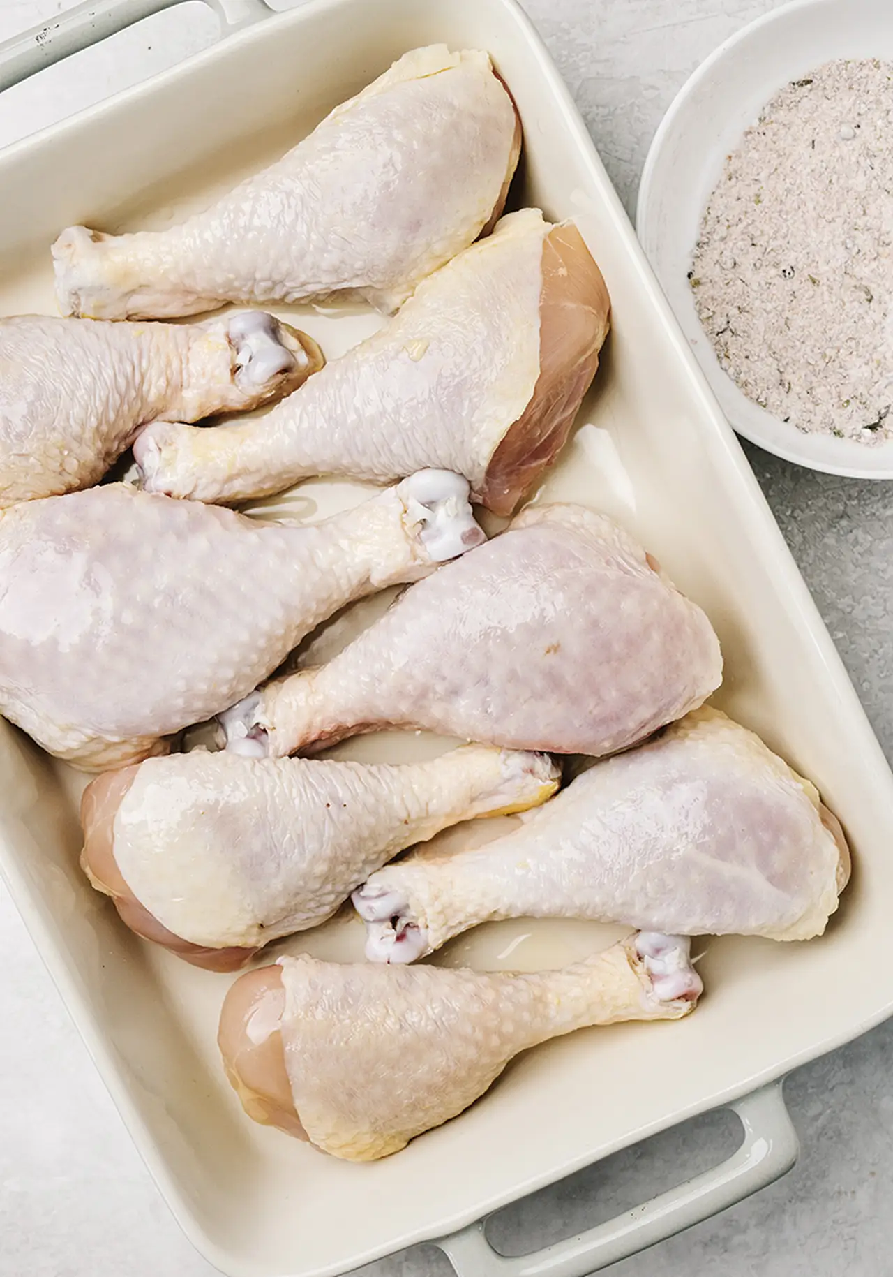 Huit pilons de poulet crus dans un plat, avec un bol de panure à côté.