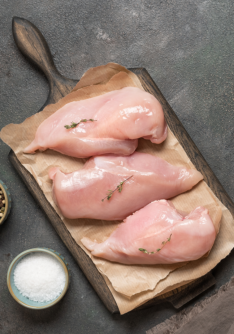 Trois poitrines de poulet crues sur une planche, avec herbes, sel et poivre.
