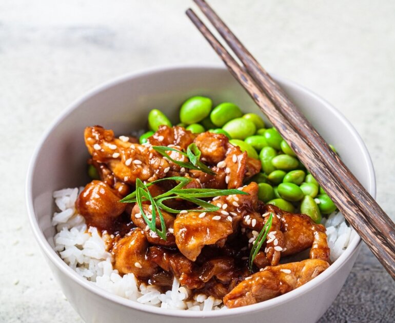 Bol de riz blanc avec poulet en sauce brune et edamame, avec des baguettes.