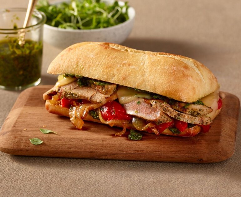 Un sandwich avec des tranches de porc, du fromage, des poivrons grillés et des légumes verts sur une planche en bois, avec un petit bol de sauce et des légumes verts à l'arrière-plan.