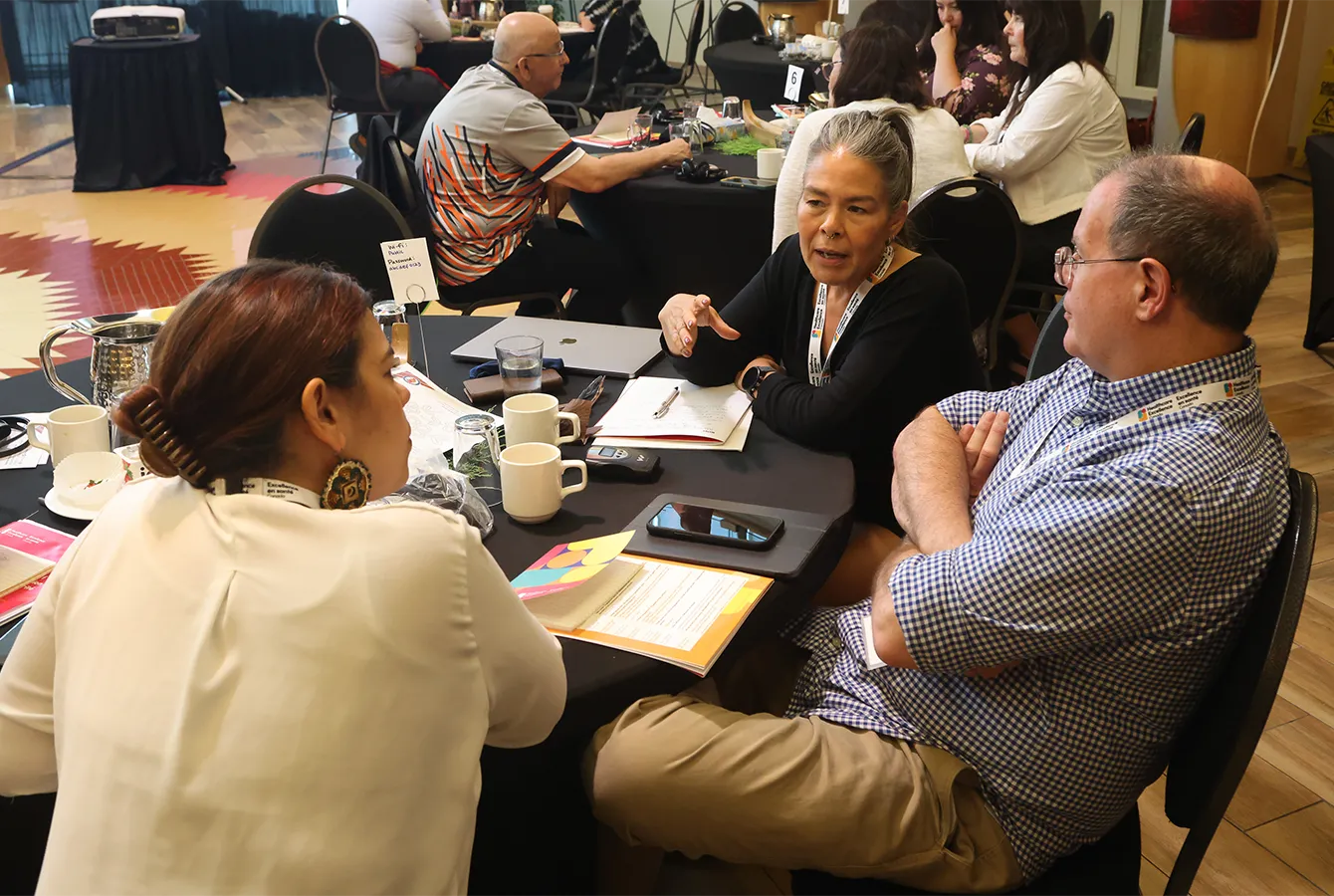 Three people sit at a round table in discussion. Papers, notebooks, and smartphones are on the table.