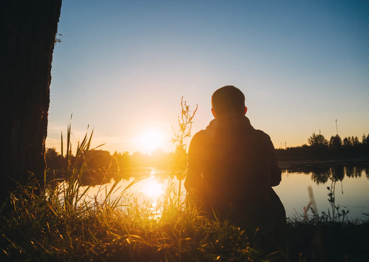 Silhouette d’une personne assise au bord d’un lac, entourée d’arbres et d’herbes, qui admire le coucher de soleil sous un ciel dégagé.