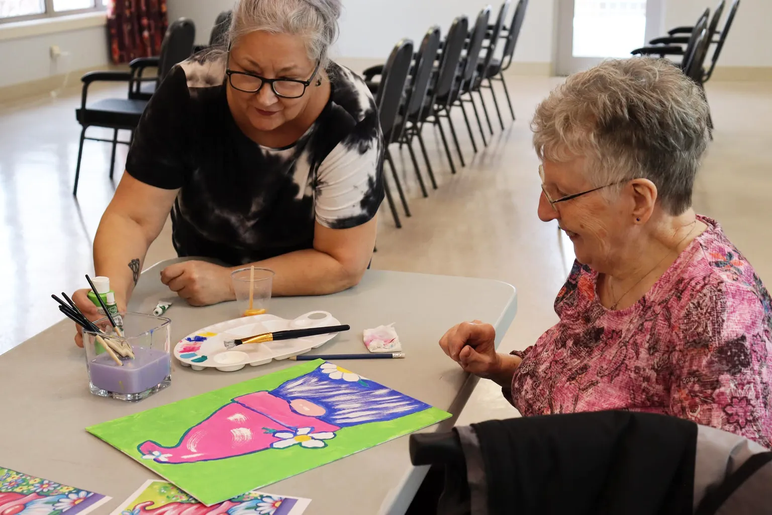 Two women sitting at a table, one painting a colorful picture while the other watches and smiles, in a bright room with chairs.
