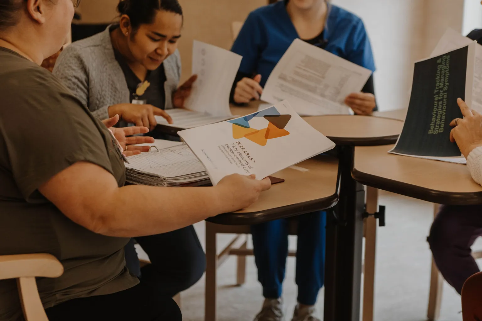 Membres du personnel de la santé assis autour de tables, examinant ensemble des documents imprimés, échangeant sur leur contenu et pointant certains éléments lors d’une séance collaborative.