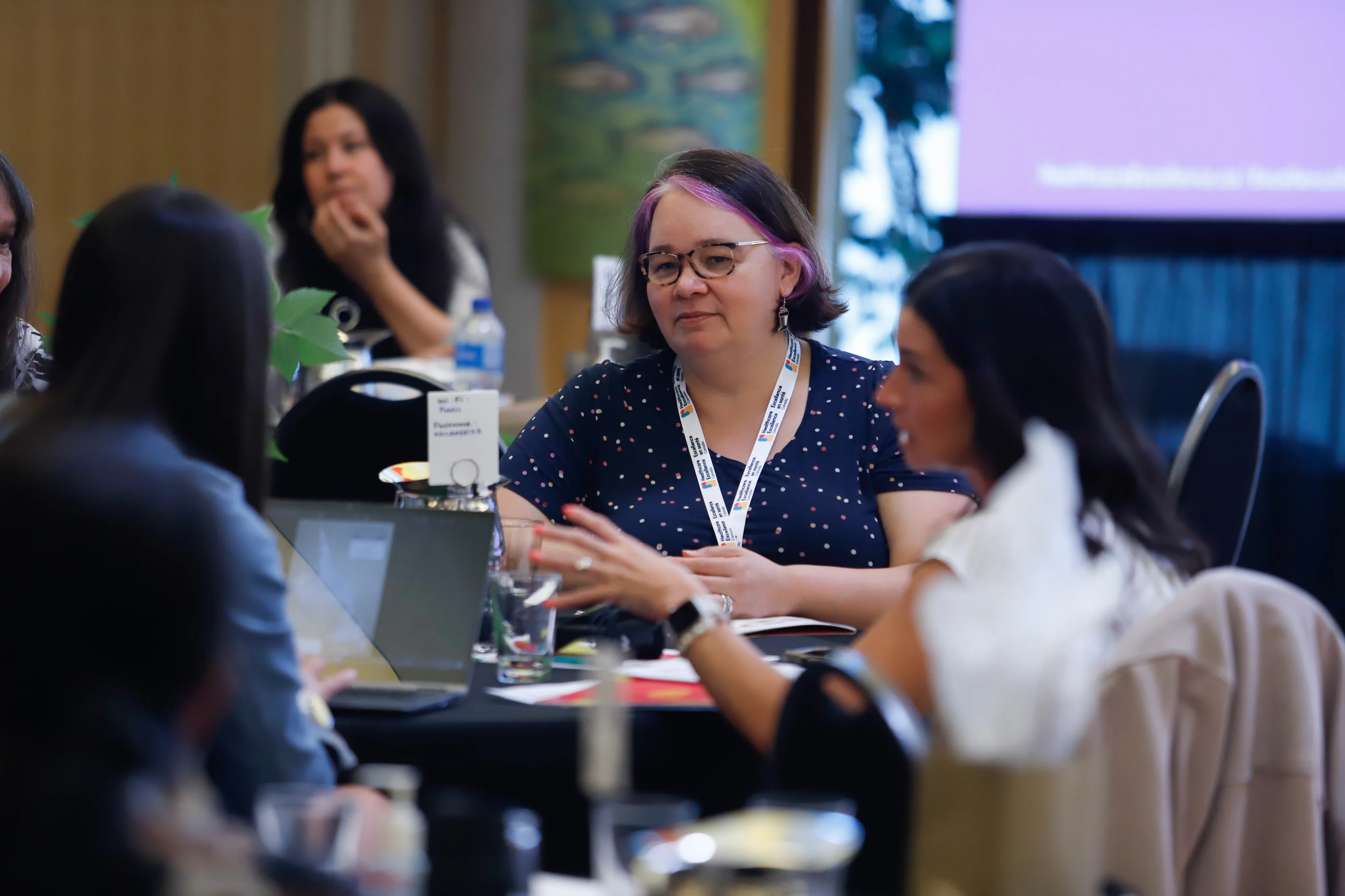 A group of people having a discussion at a conference table, with a woman in focus wearing glasses and a lanyard in a dotted shirt listening to two other people at the table.