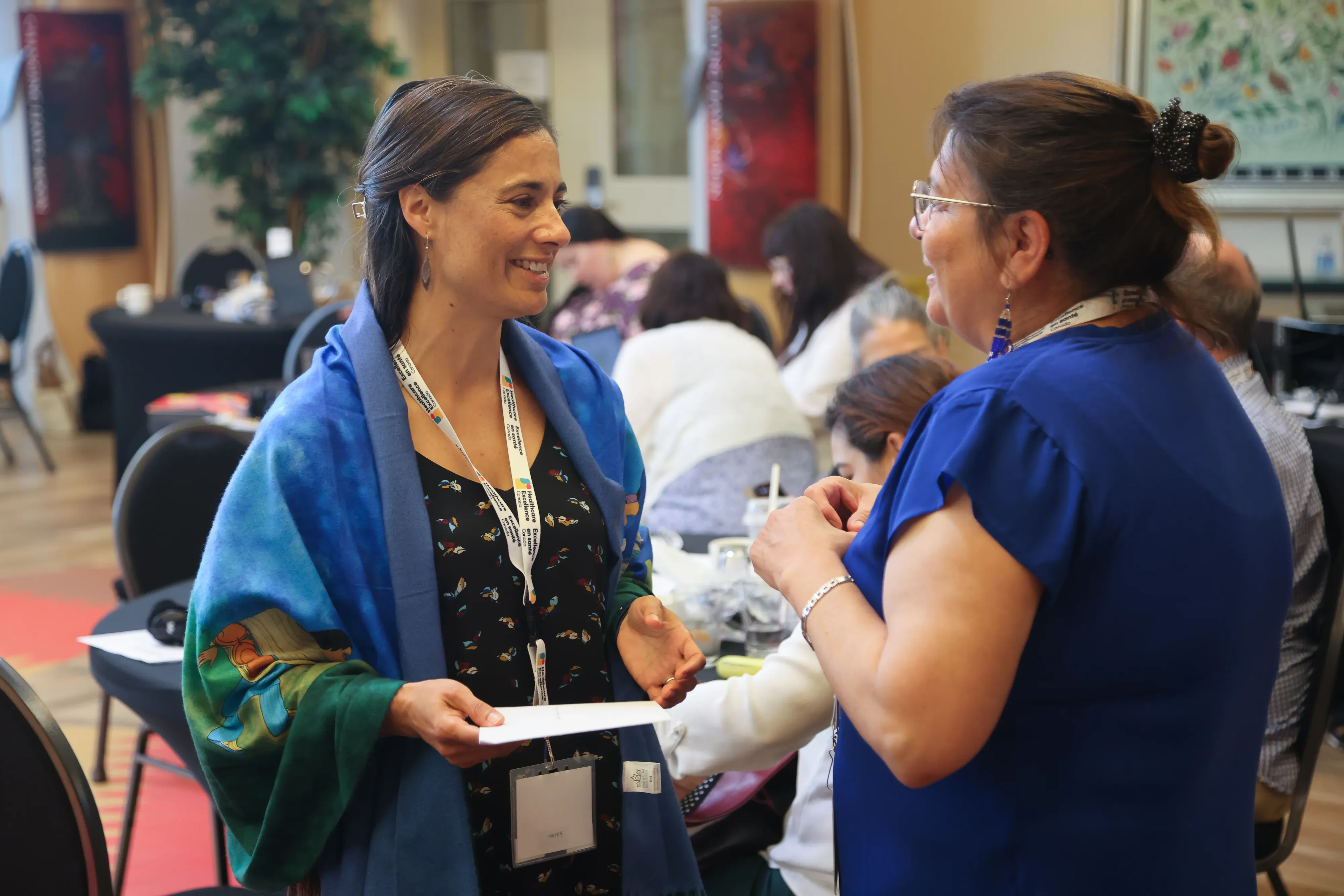 Two women are engaged in conversation at an indoor event, with people seated and interacting in the background.