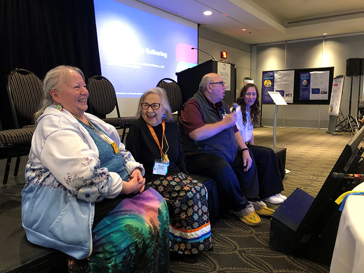 Four people sitting on a stage at a conference, with one speaking into a microphone. Presentation screen and display boards in the background.