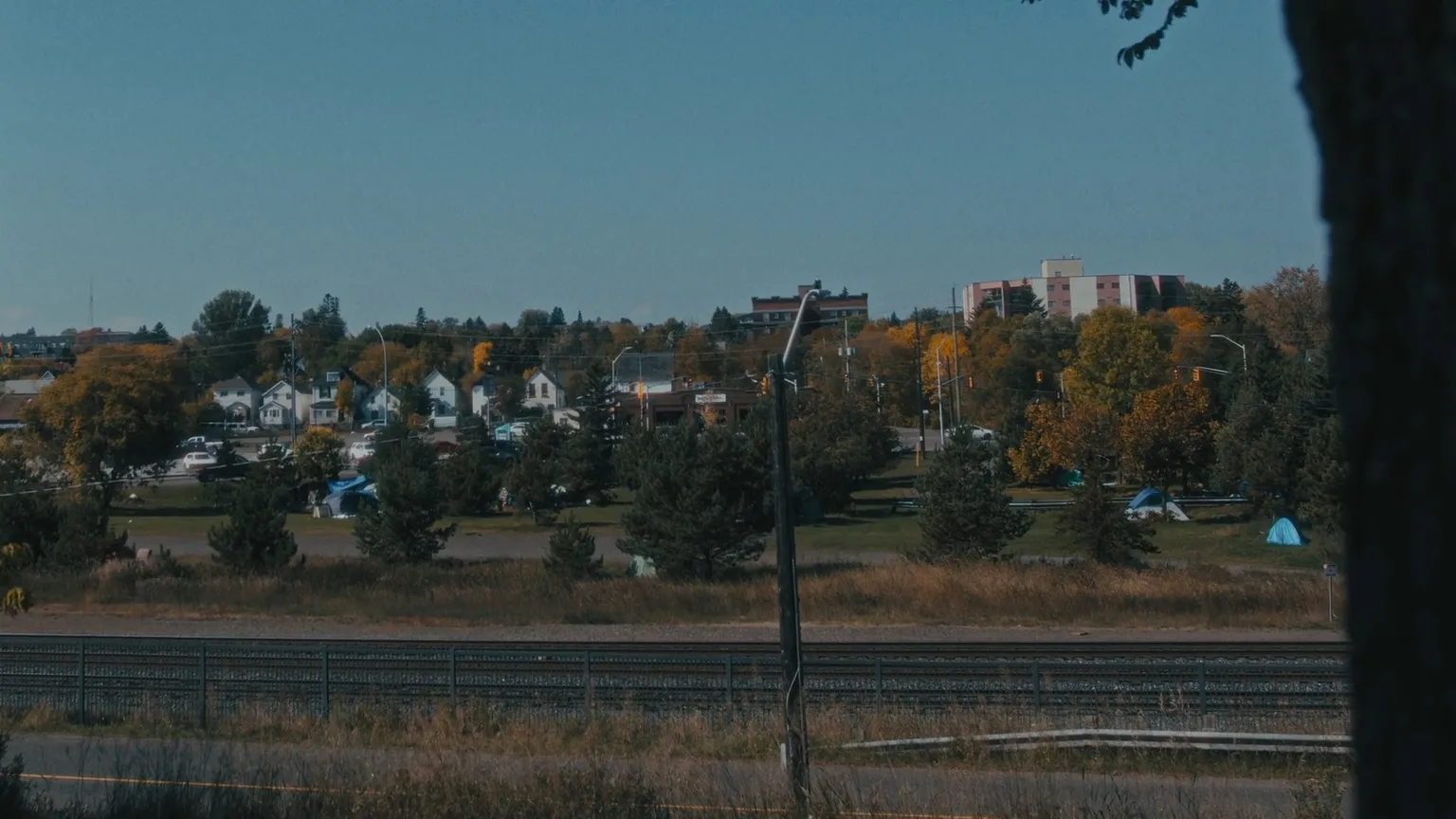 Photo showing in the background a row of houses, trees with autumn colors, and a park with tents set up. In the foreground, a railway track can be seen.